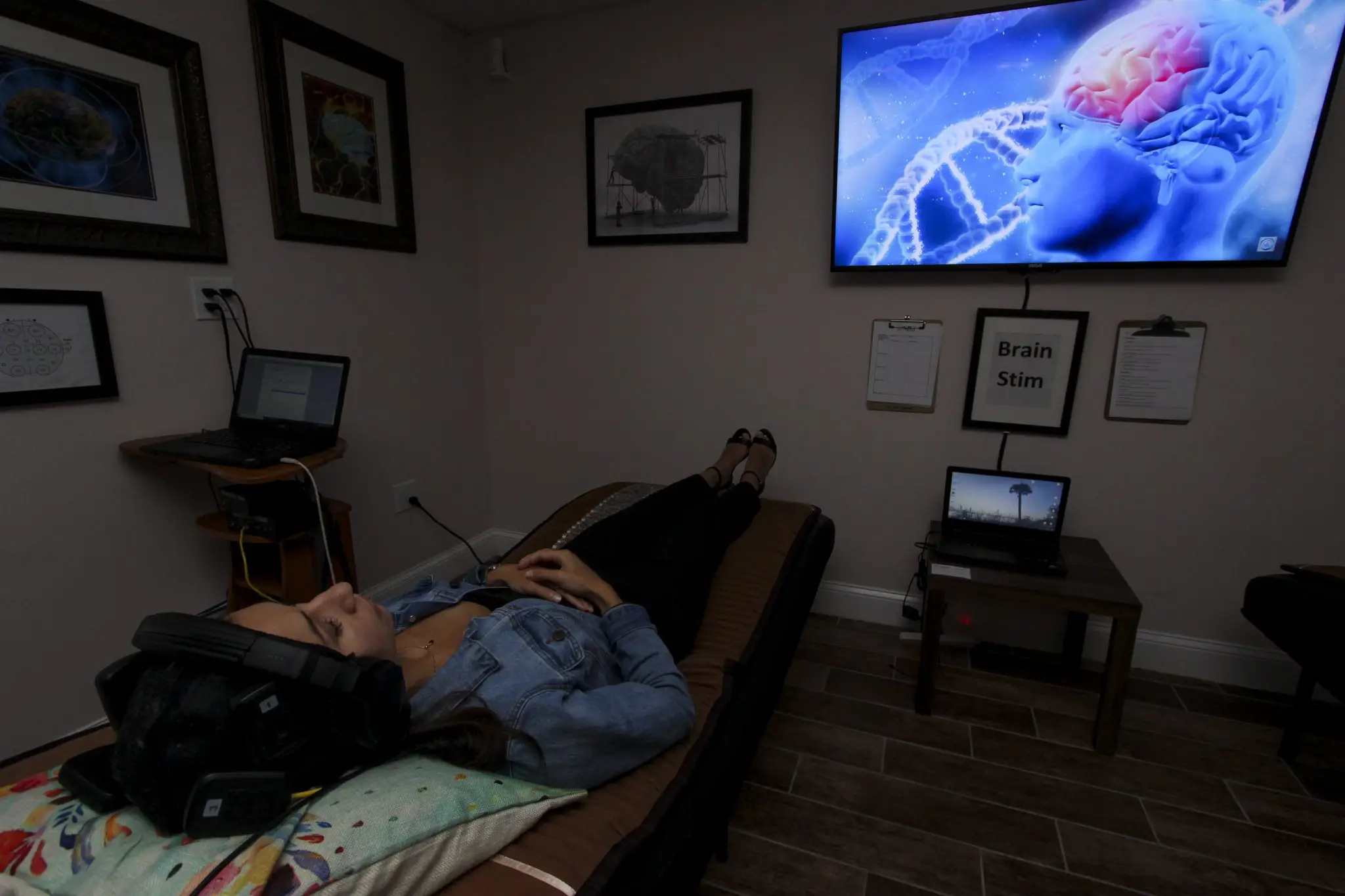 woman lying in brain stimulation chair with headphones on in dimly lit room and TV on wall showing brain graphic