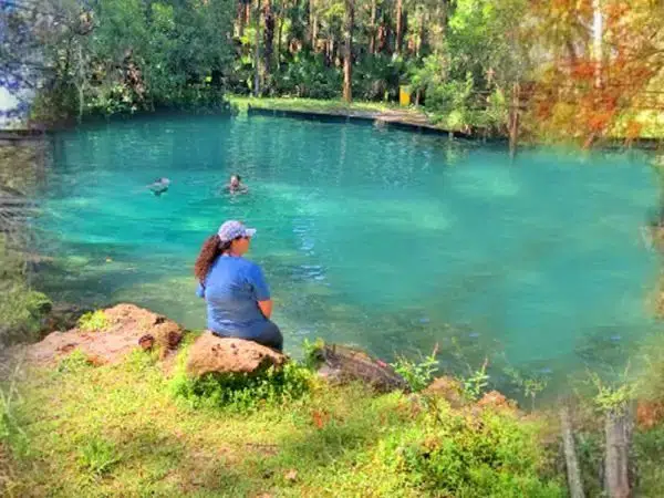 woman sitting at edge of water. people swimming in natural springs surrounded by lush greenery.