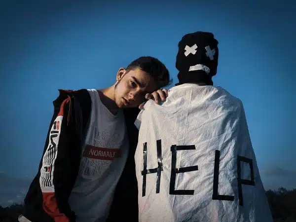 person with a blanket wrapped around them that reads HELP and another man leaning on them looking saddened.