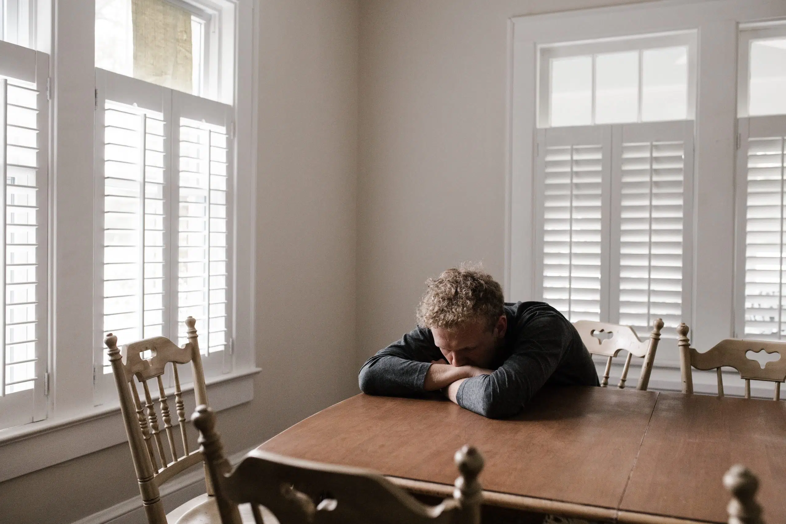 man sitting at a dining room table with his head down on his folded arms. 