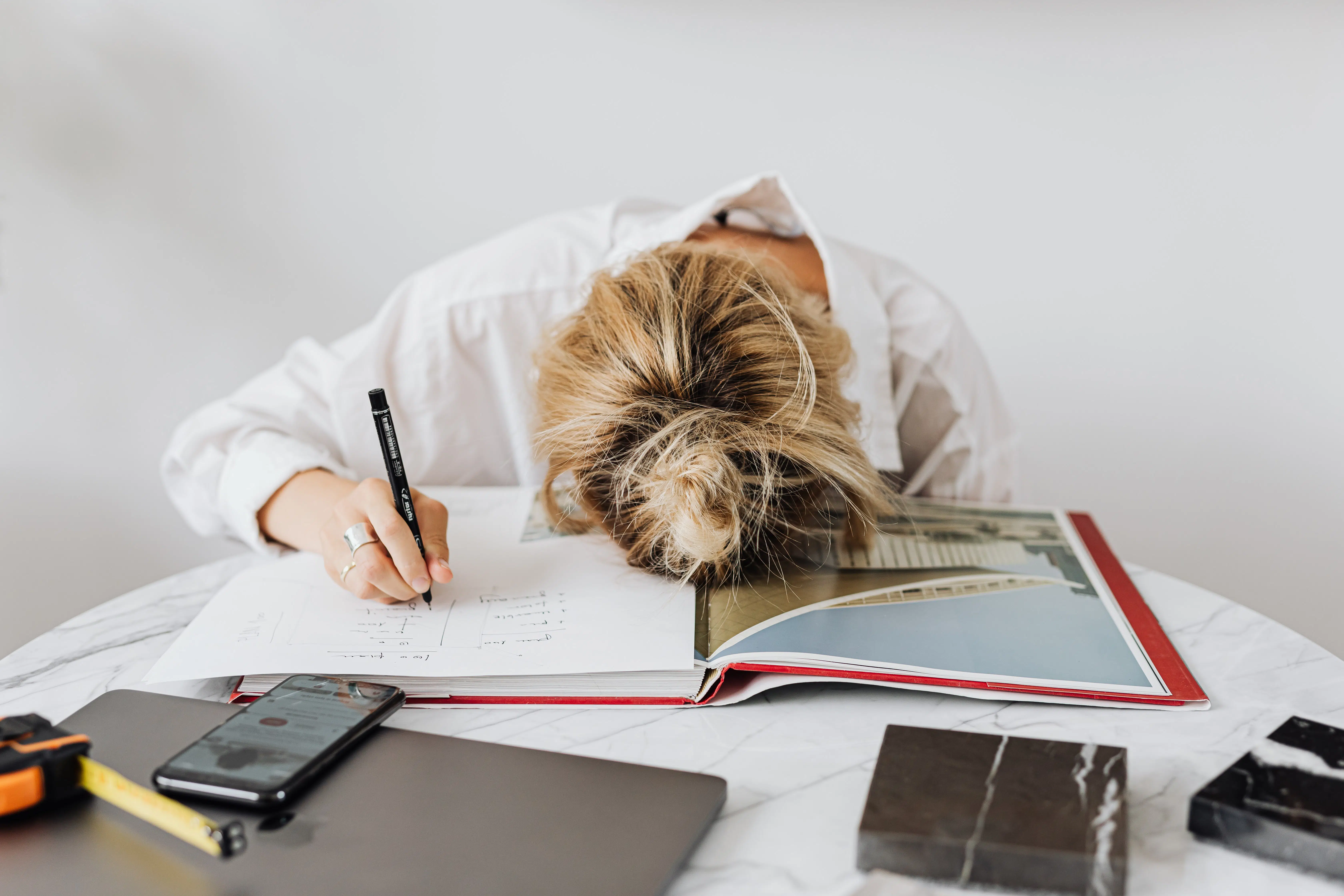 woman sitting at table with her head down, holding a pen to paper, table is covered in books, laptop, phone, measuring tape, and stone samples
