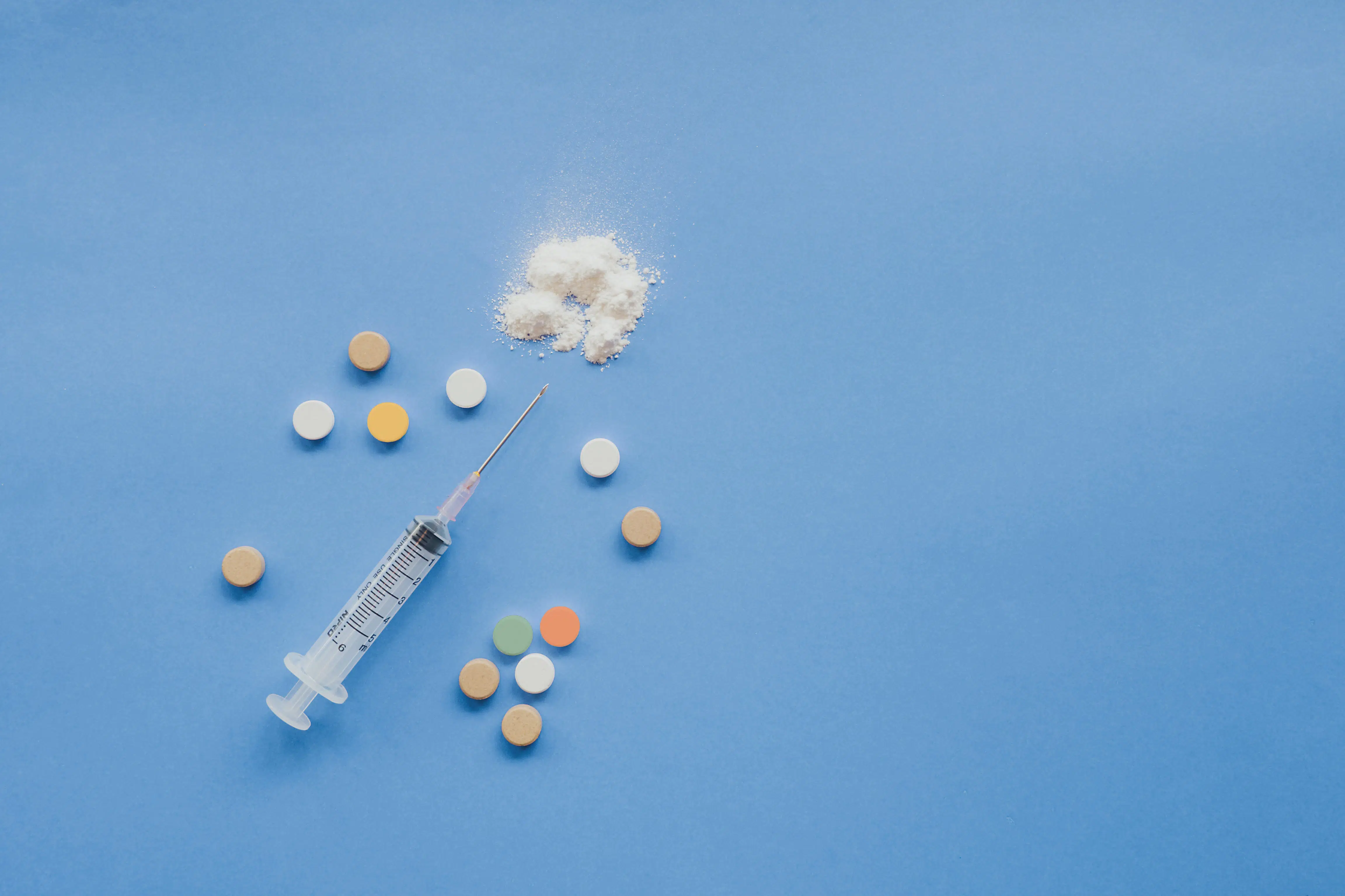 syringe, pills, and small pile of white powder displayed ona. blue background.