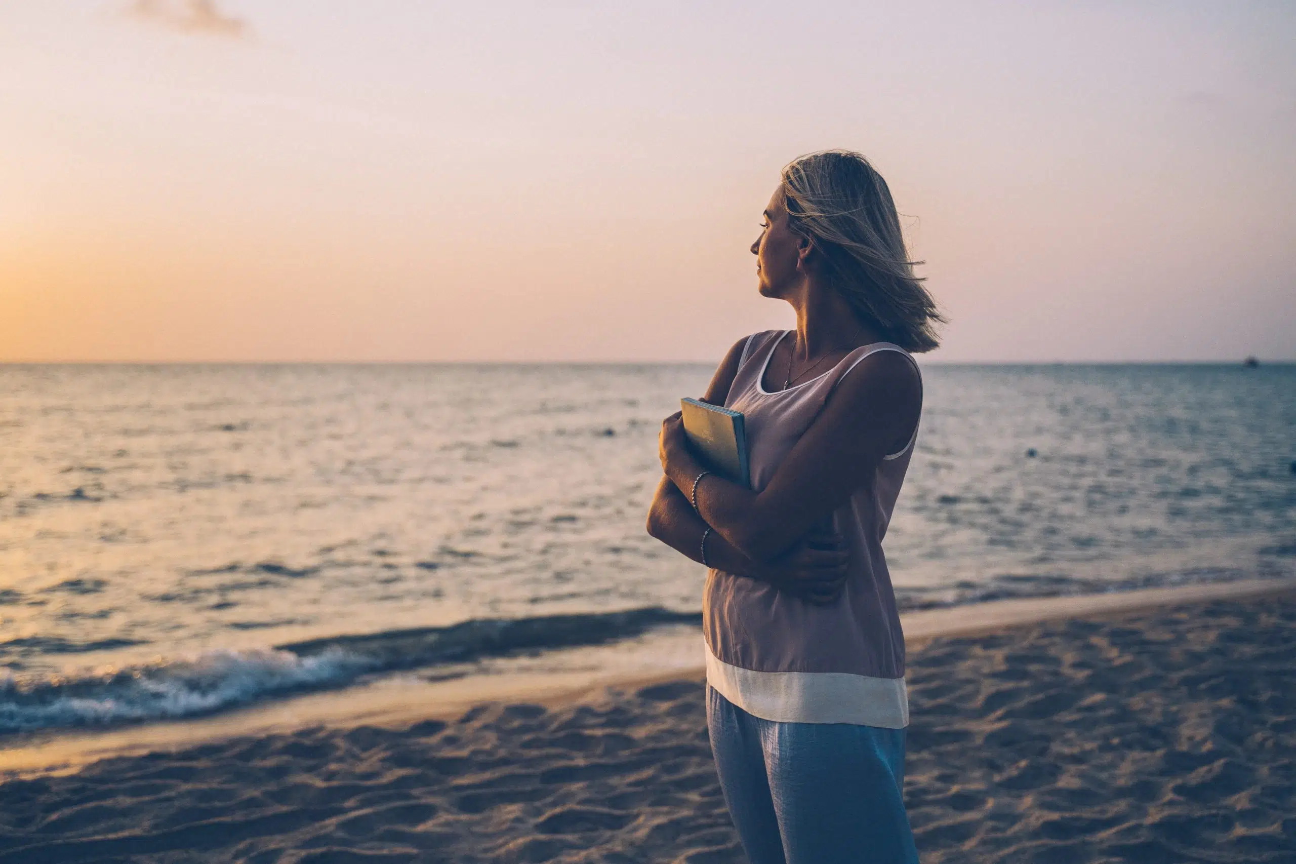 woman standing at edge of ocean, holding a book staring out into the sunset or sunrise.
