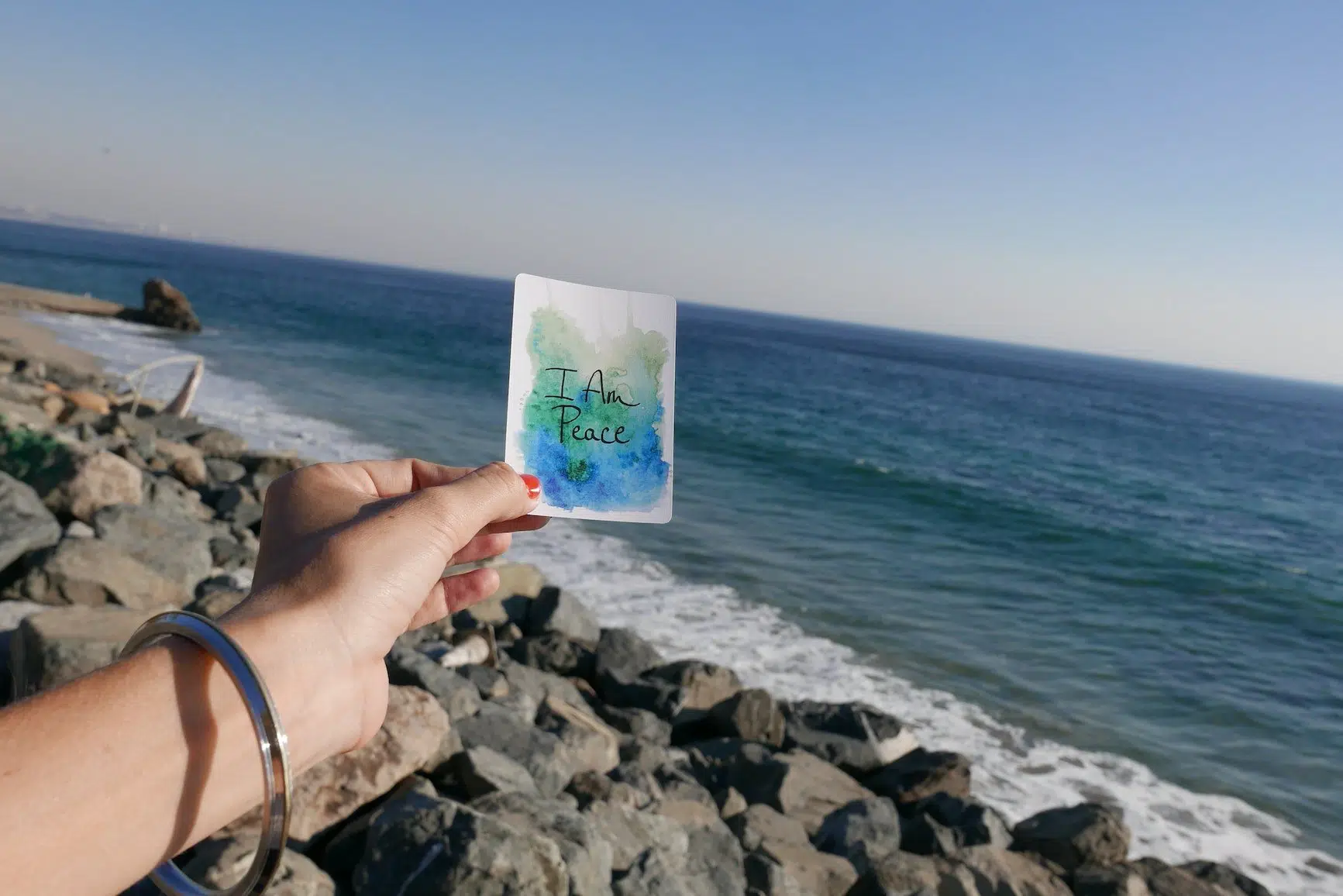woman's hand holding a small note that reads, "I am peace" with view of ocean in background.