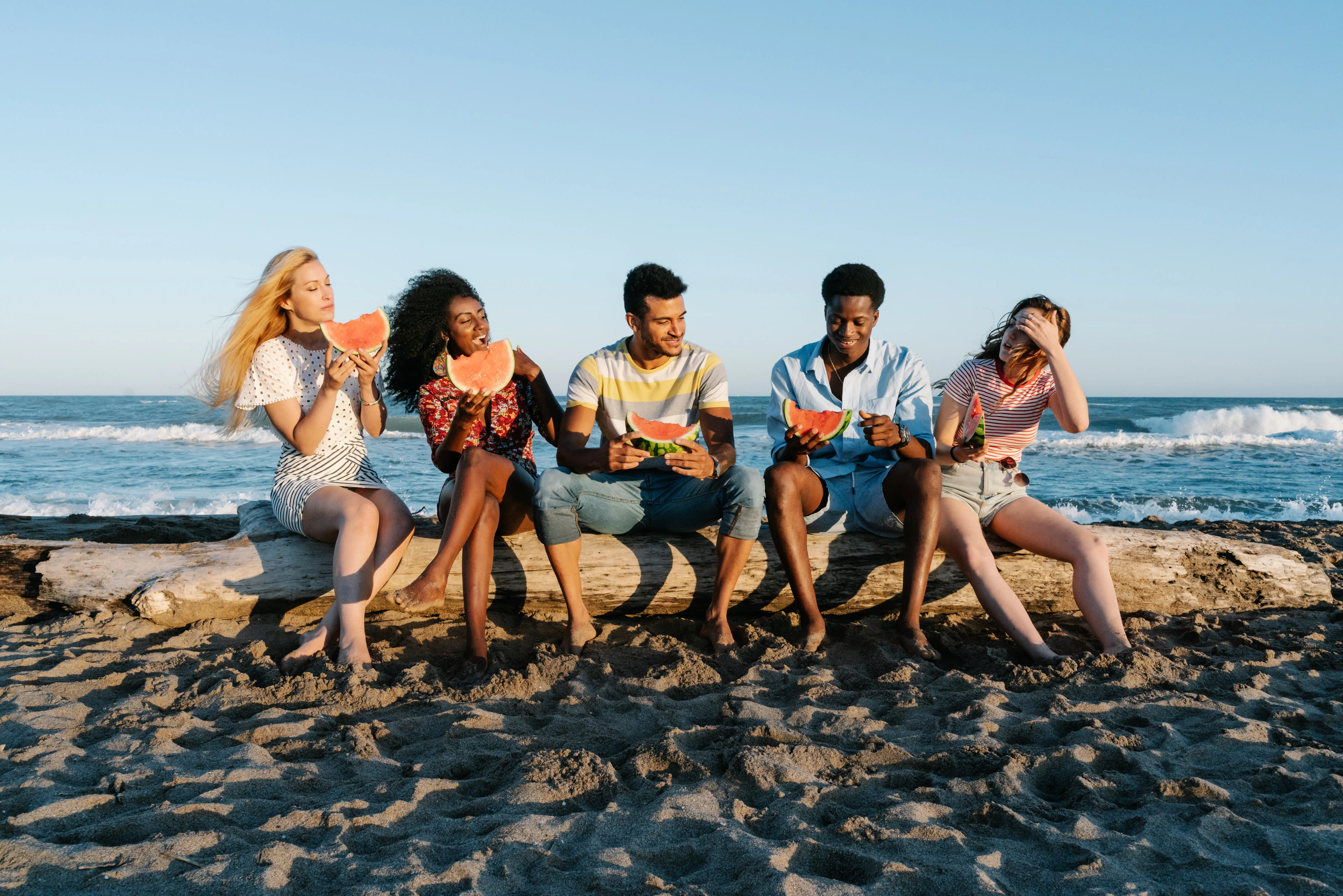 Five young adults sitting on a log on the beach eating watermelon and laughin.