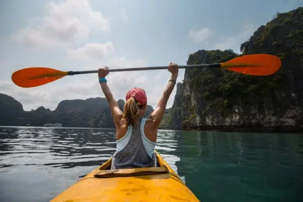 woman in kayak on the water with arms raised above head holding oar in triumphant pose.