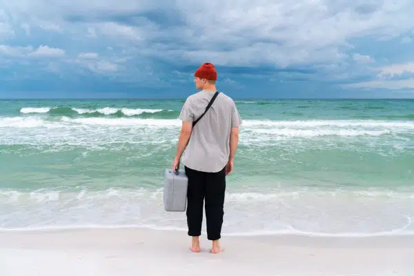 man standing at edge of ocean, staring out, with a luggage in his hand. 