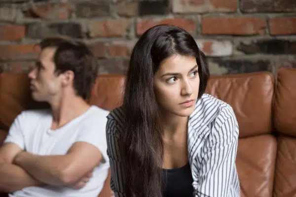 Couple sitting on couch with woman in foreground looking away, upset and concerned while man in background is looking in opposite direction, irritated and arms crossed.