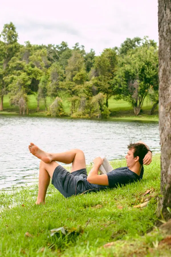 man sitting my a lake surrounded by trees, leaning back against the grass and reading a book.