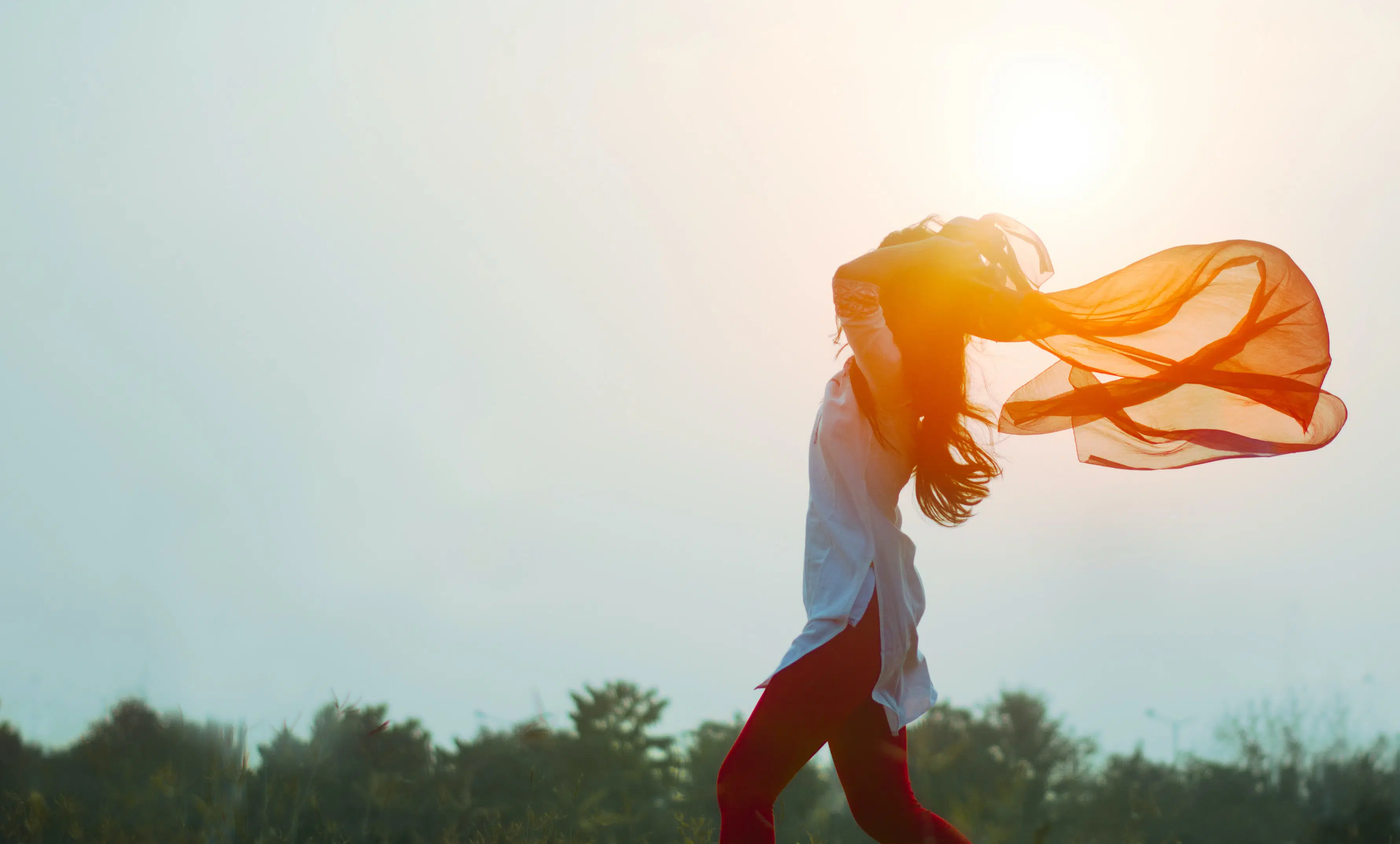 woman walking in the sun shining behind her, holding a sheer scarf blowing in the wind behind her.