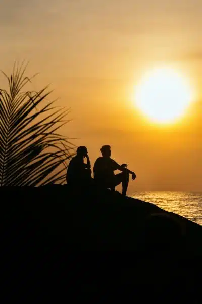 Silhouette of two people sitting on a hill with the sun setting on the ocean in the background.