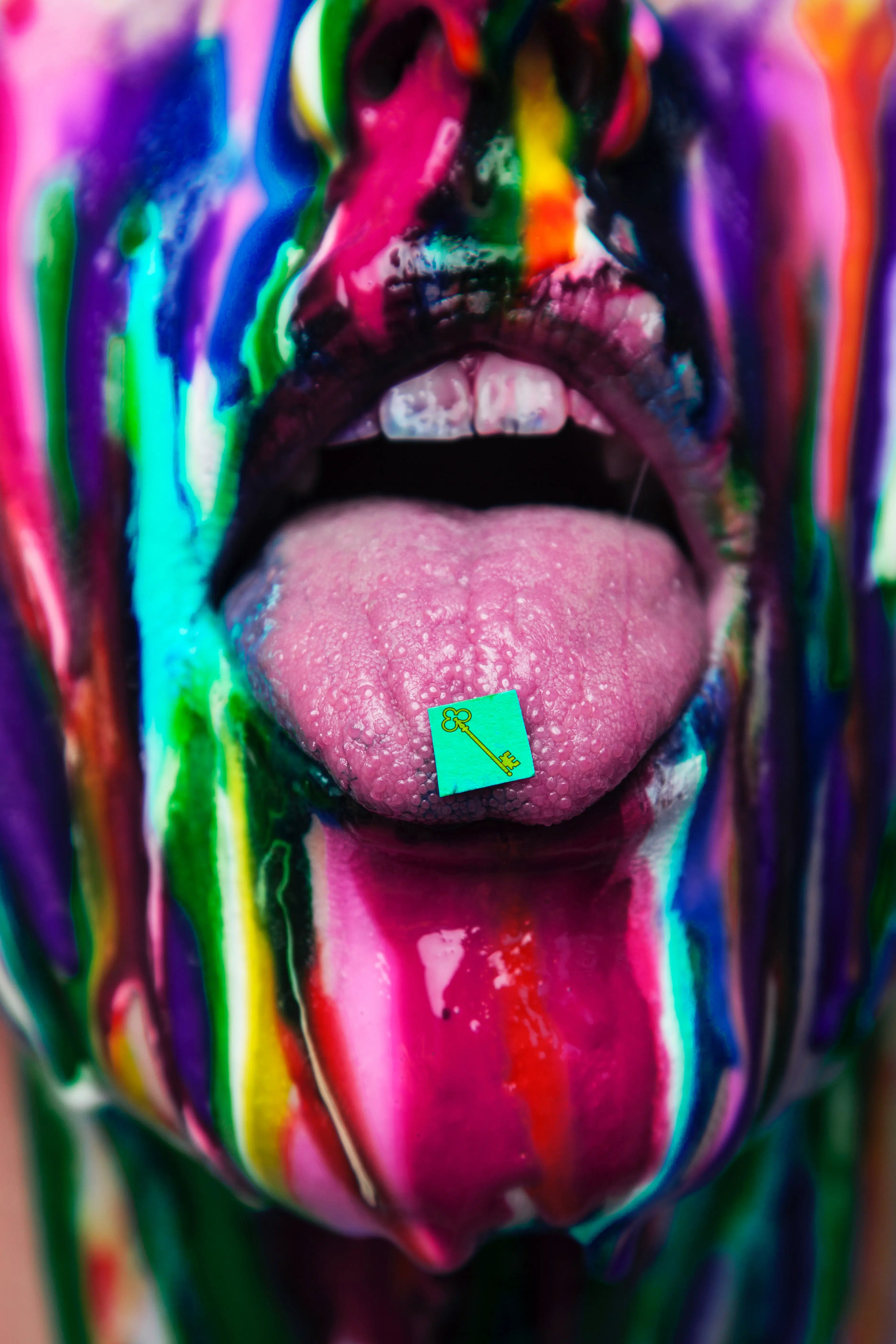 bottom half of a person's face, painted multiple colors, with their tongue sticking out and an acid tab on the tip of their tongue.