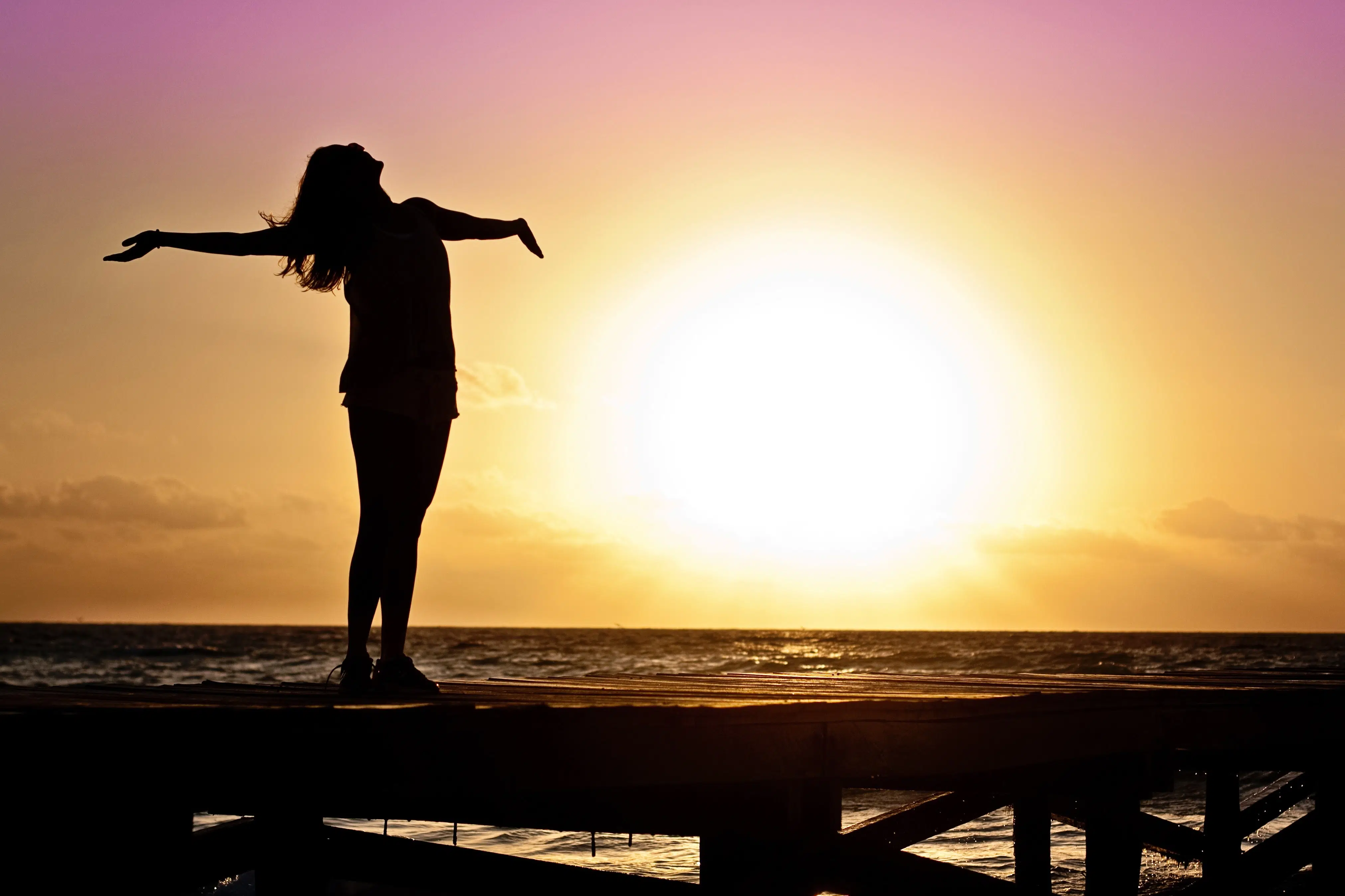 silhouette of a woman standing on ocean pier at sunrise with head leaning back and arms outstretched.