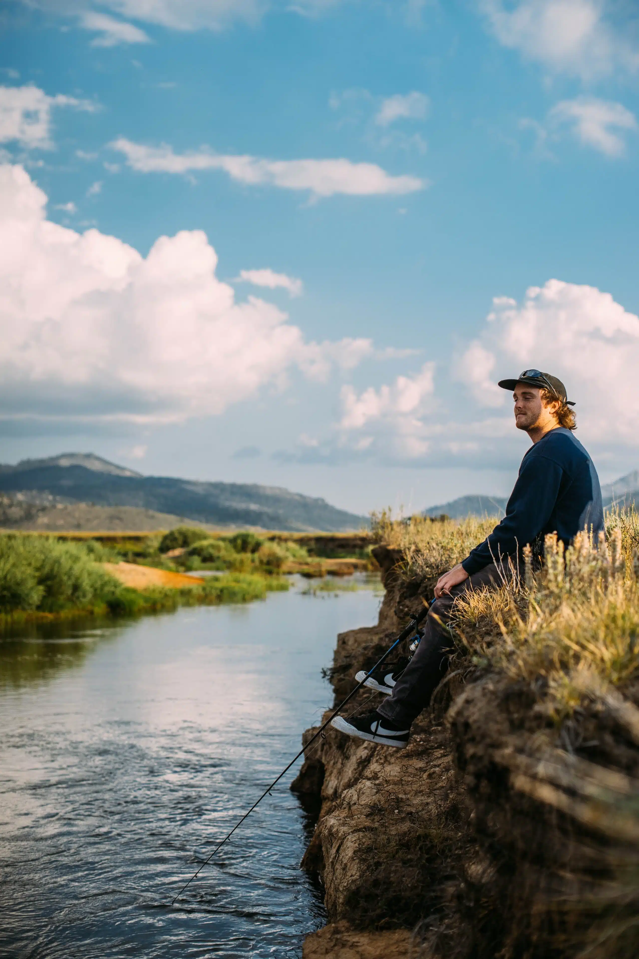 man sitting on edge of rocks fishing into stream below him, looking content.