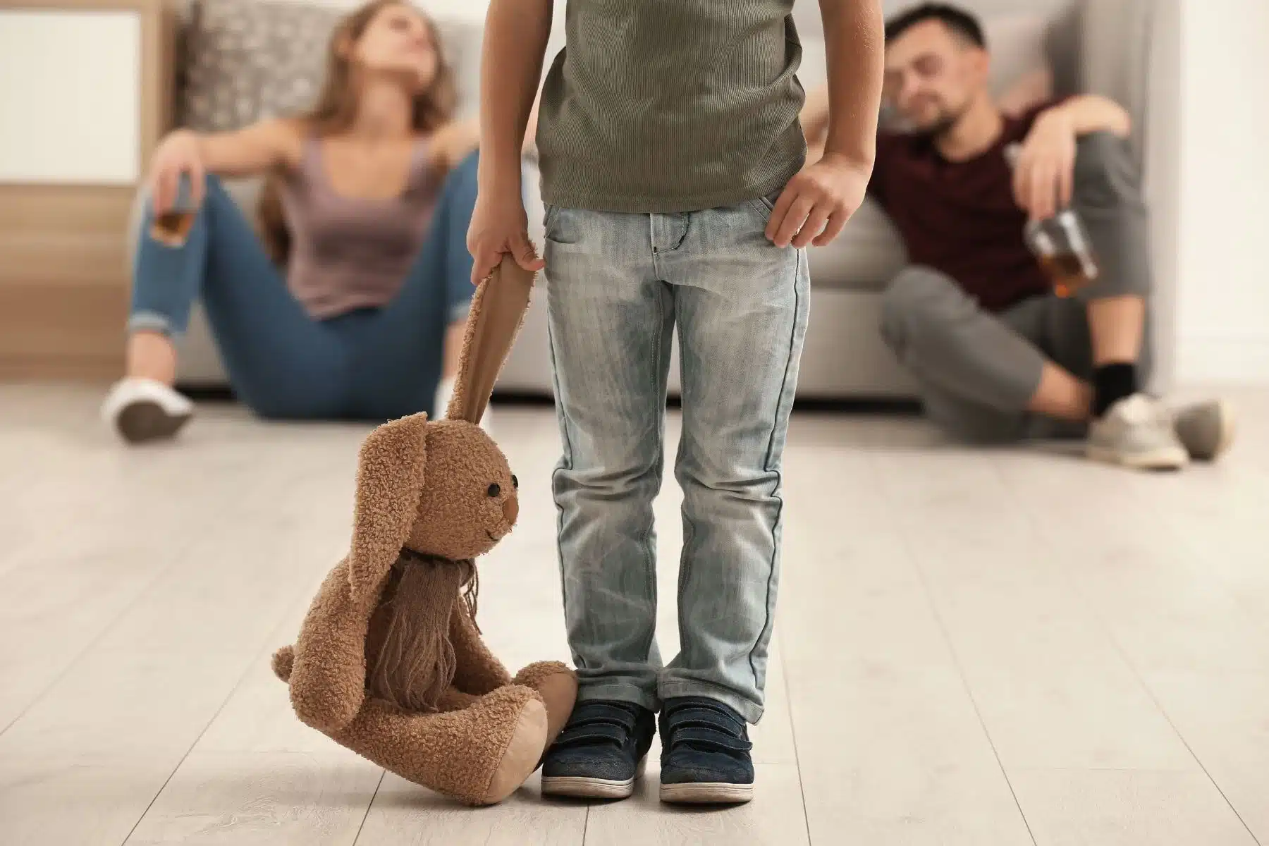 bottom half of child standing in forefront holding a worn stuffed bunny by the ear while parents are in blurred background slumped against the couch with drink in hand.